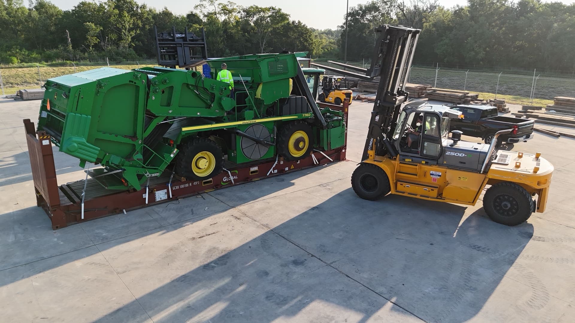 A heavy forklift alongside a John Deere cotton picker secured on a flat-rack container in the yard