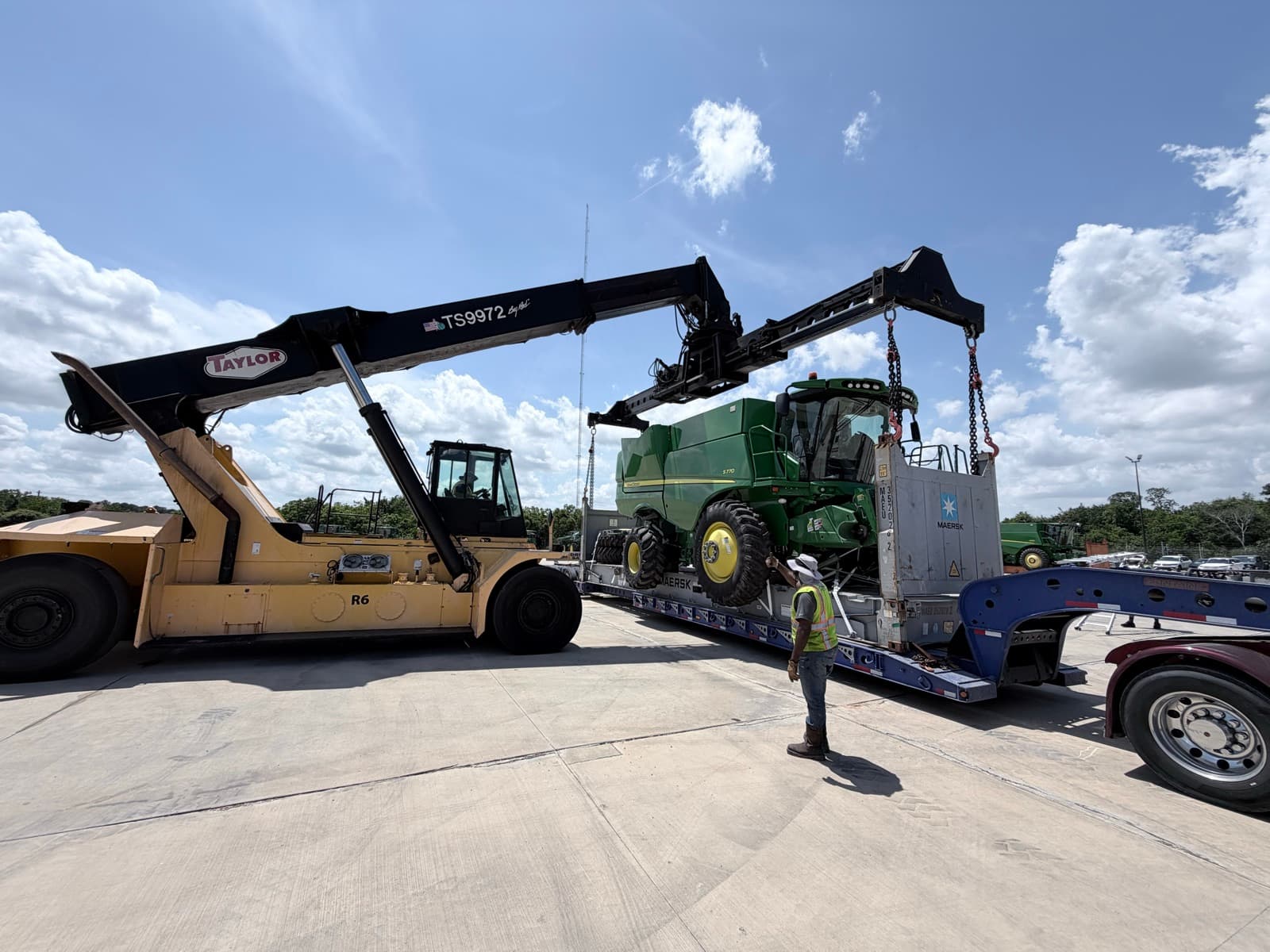 Reach stacker lifting the John Deere S770 combine over the flat-rack container before lowering