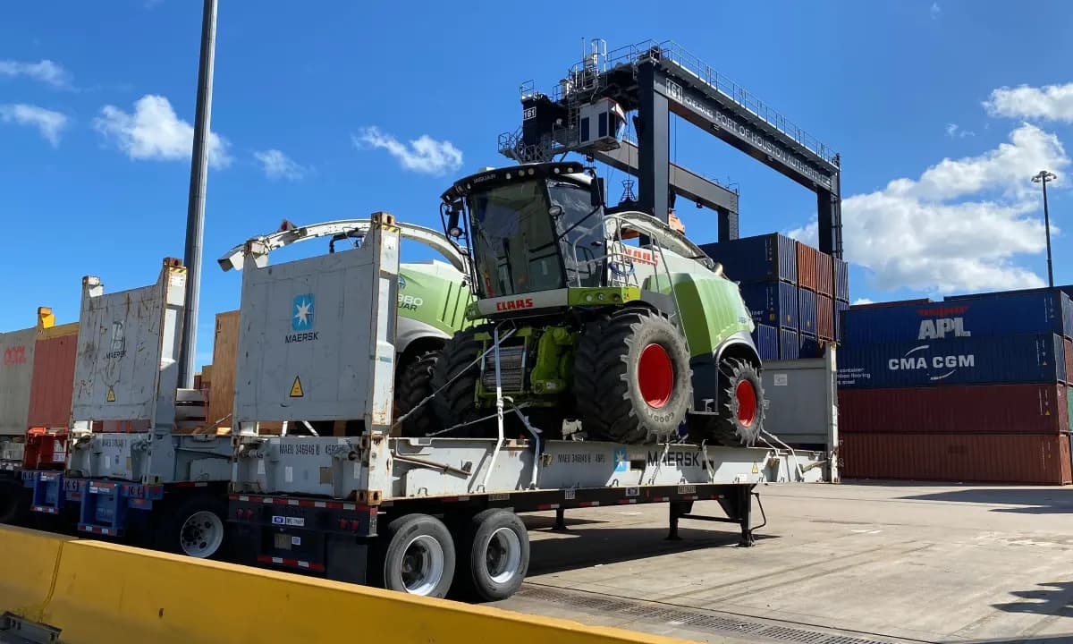 A forage harvester on a flatbed beside stacked containers and a straddle carrier.
